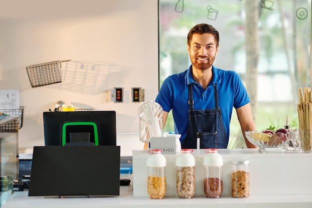 Portrait of handsome young smiling cafe worker standing at counter waiting for customers