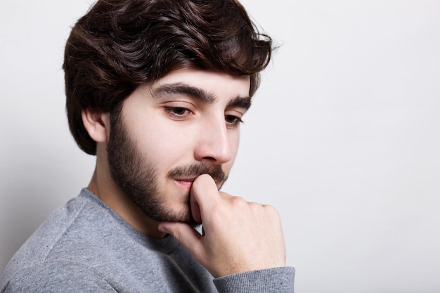 Portrait of handsome young thoughtful man with beard holding hand on chin looking down thinking about something inportant. Pensive and sad man isolated over white