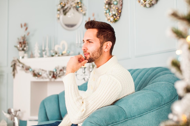 Portrait of handsome young unshaved man with stylish hairstyle wearing warm white sweater sitting in light blue armchair touching his chin and looking away Christmas mood