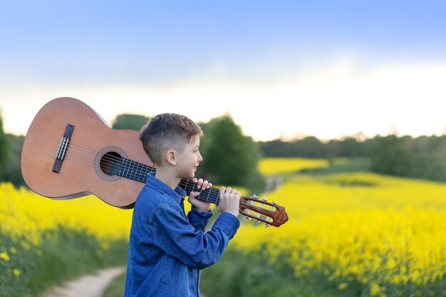 夏の黄色いフィールドを歩くギターと肖像画ハンサムボーイ。成功への道を歩む若いミュージシャン。