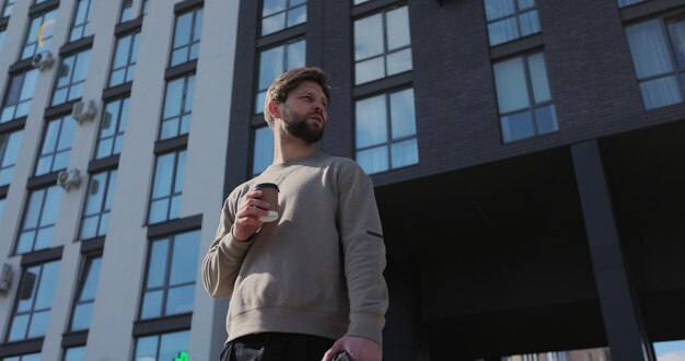 Portrait of hansome young caucasian man holding paper cup in hand at modern buildings in the background