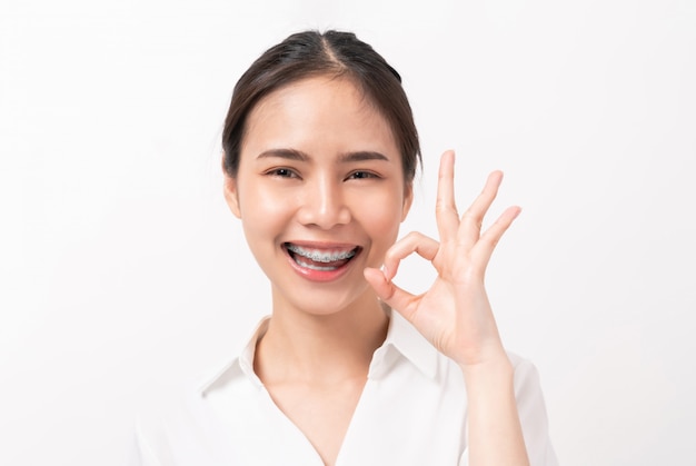 Portrait happily Asian woman shows ok sign and braces smiling on white wall,