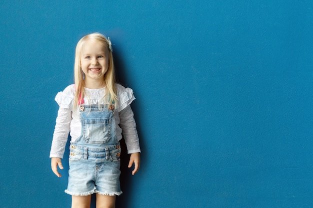 Portrait of happy 3-year-old toddler blonde girl smiling while posing against blue wall background