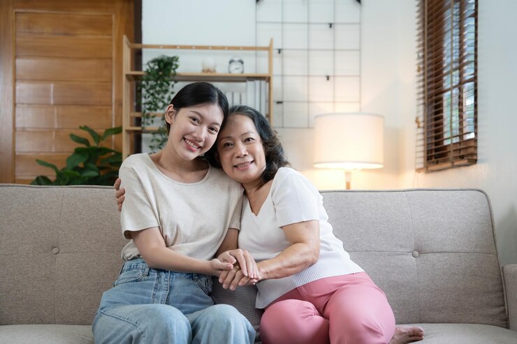 Photo 幸せな大人の娘と年配の母親が抱きしめて手をつないで自宅のソファに座っている写真若い女性と成熟した母親または祖母がカメラを見ている2世代の家族写真