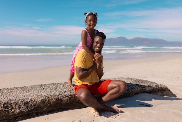 Portrait of happy african american girl embracing young father sitting on rock at beach against sky