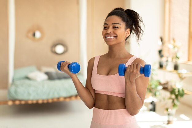 Foto ritratto di felice signora afroamericana che fa allenamento con i manubri a casa lavorando sulla forza delle braccia e sorridente copia spazio