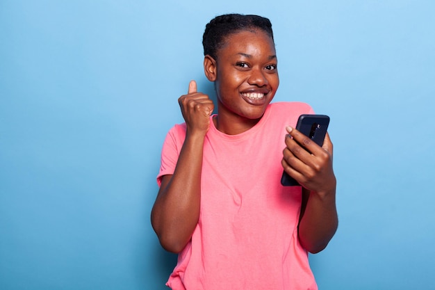 Portrait of happy african american teenager received a test message from friend enjoying good news standing in studio with blue background. Young woman using smartphone browsing on social media