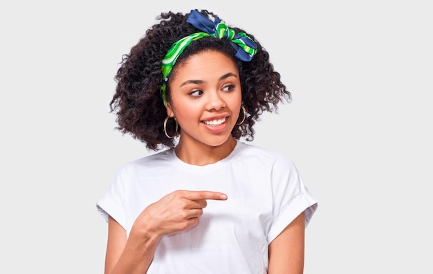 Portrait of happy African American young woman dressed in white t shirt and green trendy headband indicates aside with forefinger to blank copy space isolated over white wall