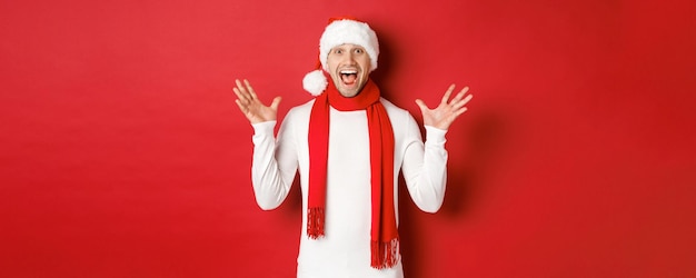 Portrait of happy and amazed handsome man celebrating new year wishing merry christmas wearing santa...