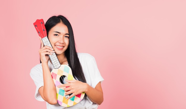 Portrait of happy Asian beautiful young woman teen confident smiling face hold acoustic Ukulele guitar, female playing Hawaiian small guitar, studio shot isolated on pink background, with copy space