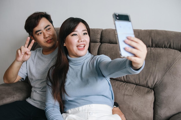 Portrait of happy asian couple sitting on sofa and taking selfie together by smartphone at home