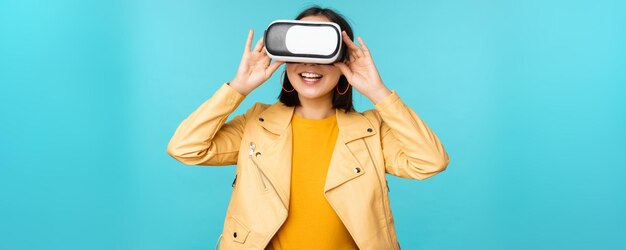 Portrait of happy asian female model using VR headset smiling and laughing in virtual reality glasses standing over blue background