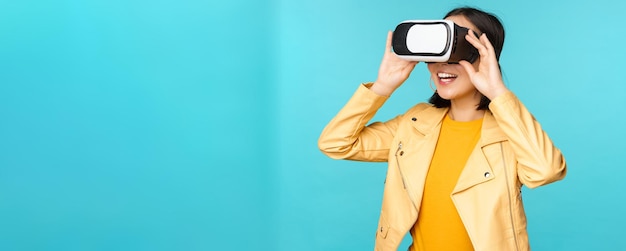 Portrait of happy asian female model using VR headset smiling and laughing in virtual reality glasses standing over blue background