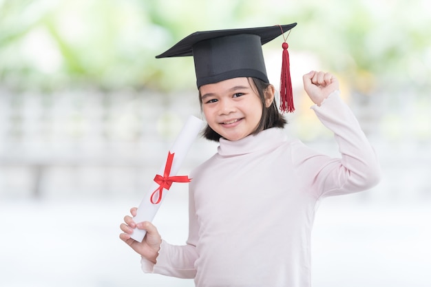 Portrait happy Asian female school kid graduate in a graduation cap holds a rolled certificate. Graduation Celebration Concept Stock Photo