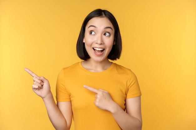 Портрет счастливой азиатской девушки, указывающей пальцами и смотрящей влево, улыбающейся, изумленно проверяющей промо-баннер с рекламой на желтом фоне
