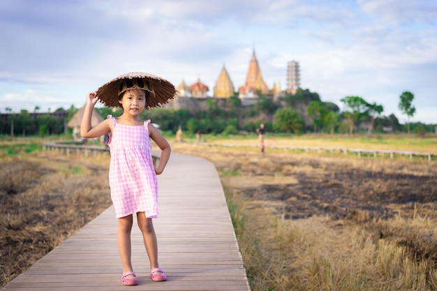 幸せなアジアの少女の肖像画は、寺院と橋の歩道に立っているドレスで帽子を着用します。