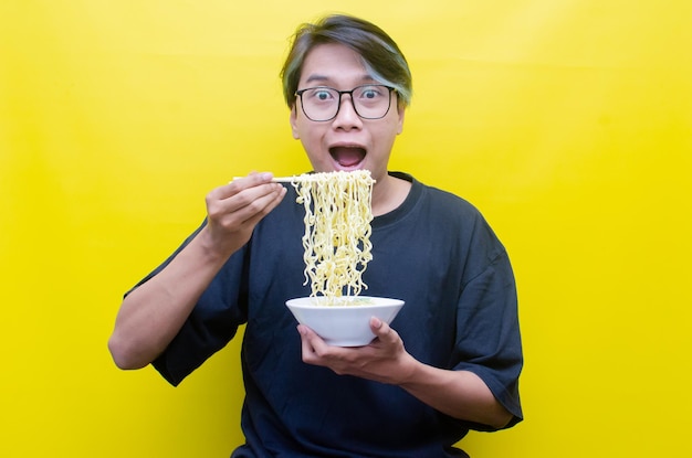 Портрет счастливого азиата в черной футболке ест лапшу быстрого приготовления с помощью палочек для еды и изолированной миски