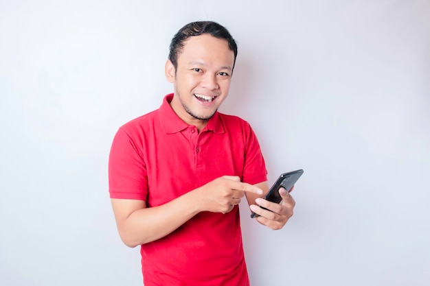 A portrait of a happy Asian man is smiling and holding his smartphone wearing a red tshirt isolated by a white background