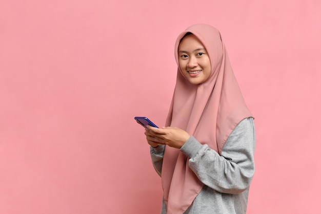 Portrait happy asian muslim woman feeling happiness and looking camera holding smartphone on pink background. Cute asian muslim girl smiling wearing casual hijab and connect internet shopping online