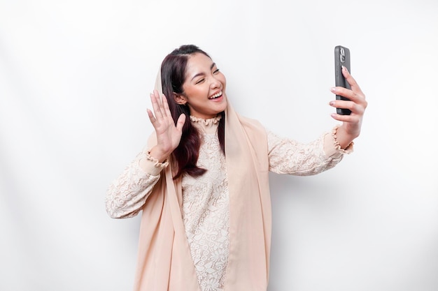 A portrait of a happy Asian Muslim woman wearing a headscarf holding her phone isolated by white background