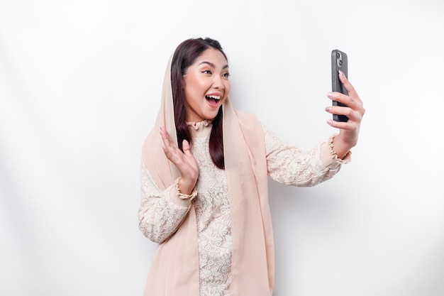 A portrait of a happy Asian Muslim woman wearing a headscarf holding her phone isolated by white background