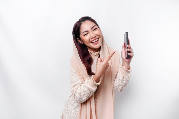 A portrait of a happy Asian Muslim woman wearing a headscarf holding her phone isolated by white background