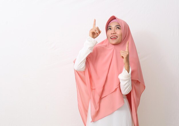 A portrait of happy asian muslim woman wearing a veil or hijab\
pointing or presenting something above her. isolated on white\
background with copy space