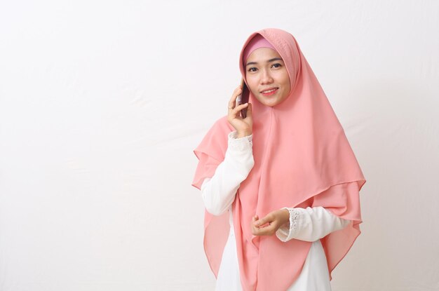 A portrait of happy asian muslim woman wearing a veil or hijab\
while speaking on the phone, having a nice conversation with\
someone. isolated on white background with copy space