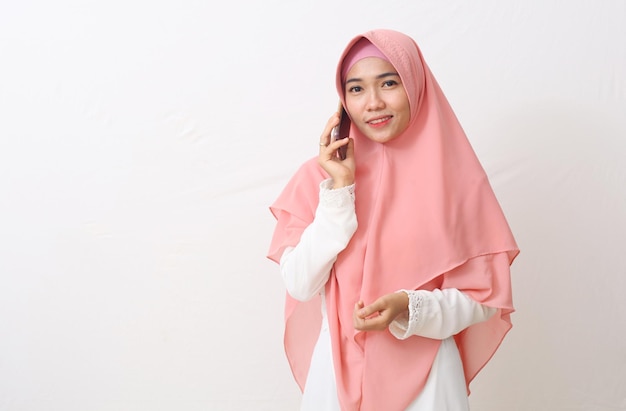 A portrait of happy asian muslim woman wearing a veil or hijab\
while speaking on the phone, having a nice conversation with\
someone. isolated on white background with copy space