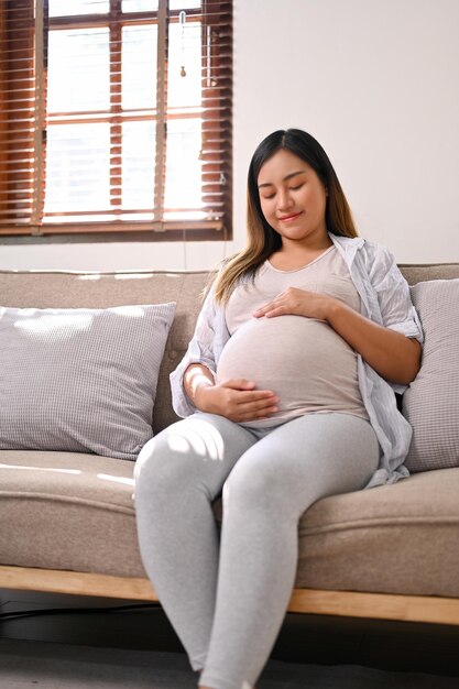 Il ritratto della donna incinta asiatica felice si rilassa sul sofà nel suo salotto