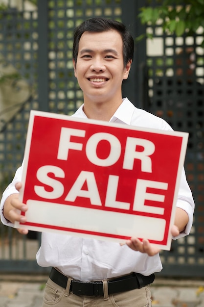 Портрет счастливого азиатского агента по недвижимости, показывающего вывеску на продажу