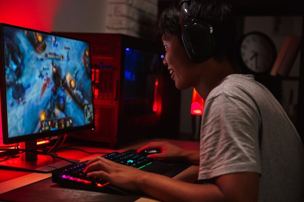 Portrait of happy asian teenage gamer boy playing video games on computer in dark room, wearing headphones and using backlit colorful keyboard