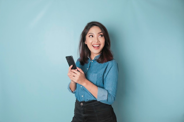 행복한 아시아 여성의 초상화가 웃고 있고 파란색 배경에 격리된 파란색 셔츠를 입고 스마트폰을 들고 있다