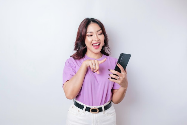 행복한 아시아 여성의 초상화가 흰색 배경에 격리된 라일락 보라색 티셔츠를 입고 웃고 있고 스마트폰을 들고 있다