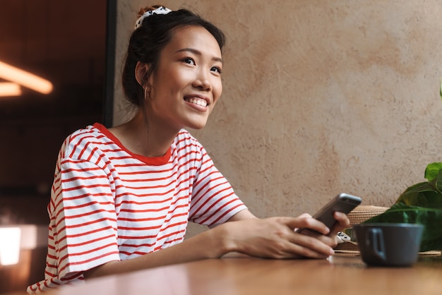 Портрет счастливой азиатской женщины, улыбающейся и держащей мобильный телефон, сидя в кафе в помещении