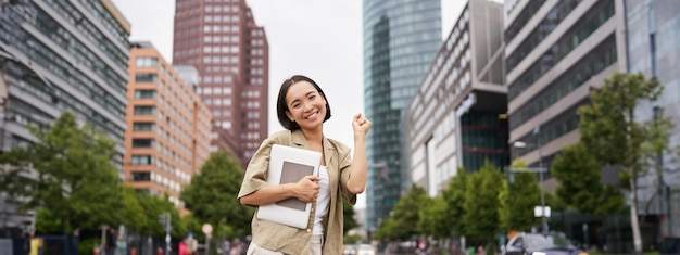 幸せなアジアの女性の肖像画は、通りの道路の近くにタブレットを持って立ち、手を上げて応援しています