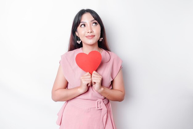 A portrait of a happy Asian woman wearing a pink blouse holding a red heartshaped paper isolated by white background