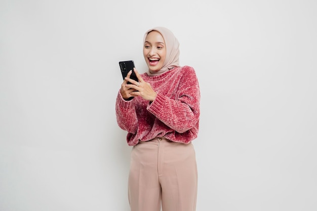 A portrait of a happy Asian woman wearing a pink sweater and hijab holding her phone isolated by white background