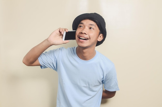 Portrait of happy asian young man wearing black hat while on\
the phone isolated background