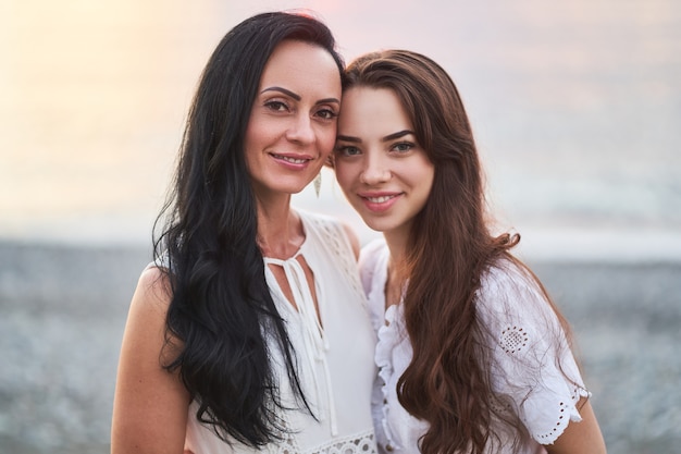 Portrait of happy attractive mother with young daughter looking together at a camera in summertime at sunset by the sea