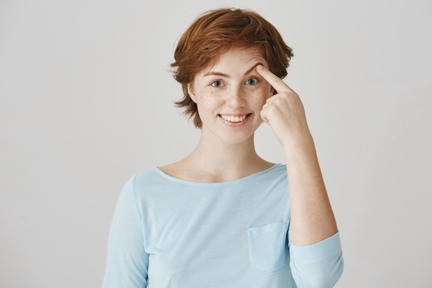 Portrait of happy attractive redhead female student with freckles in ordinary clothes pulling brow with index finger smiling and being in good mood