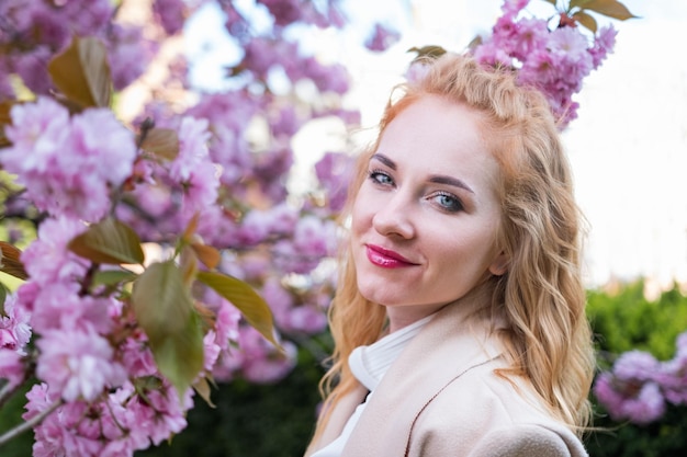 Foto ritratto di una donna felice e attraente con un cappotto beige tra i rami di sakura in fiore una ragazza elegante con le labbra rosse in un parco in fiore