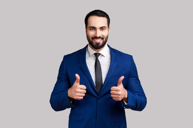 Portrait of happy bearded businessman showing thumbs up and smiling excitedly, enjoying excellent result, wearing official style suit. Indoor studio shot isolated on gray background.