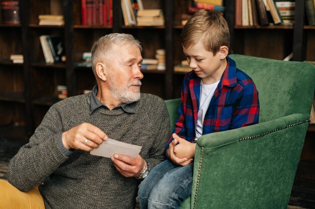 Ritratto di felice nonno barbuto che parla con il nipote carino, divertendosi a guardare un vecchio album fotografico, godendosi i ricordi guardando l'album di foto di famiglia a casa in un'accogliente stanza sullo sfondo degli scaffali