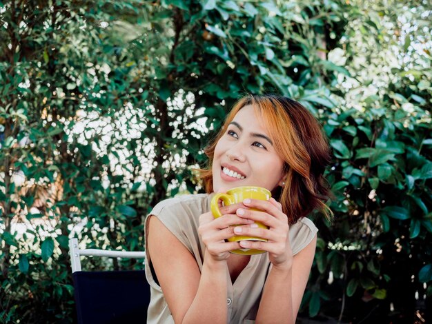 Portrait of a happy beautiful adult Asian woman with a highlight trendy short hairstyle drinking coffee holding the yellow mug looking up and sitting in the green garden background