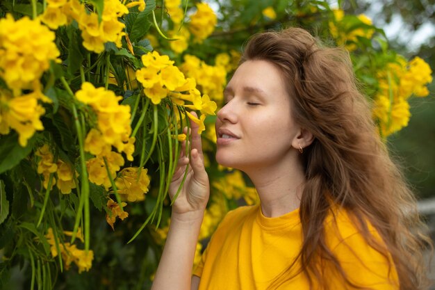 ひげを生やした幸せな美しいひげを生やした女の子の若いポジティブな女性の肖像画は、庭で美しい黄色の花の匂いを嗅いでおり、春や夏の日を楽しんで、深く新鮮な空気を呼吸しています