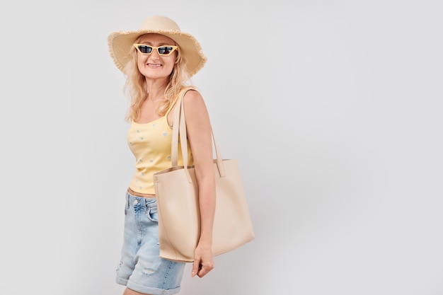 Portrait of happy beautiful blonde mature woman in yellow summer clothes straw hat sunglasses and shopping bag smiling isolated on white studio background Vacation and travel concept