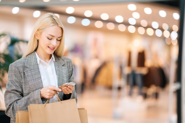 Portrait of happy beautiful blonde young woman in stylish clothes using mobile phone holding shopping paper bags with purchases in mall