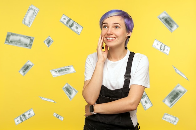 Portrait of happy beautiful hipster girl with violet short hair\
in denim overalls holding palm near face and dreaming about many\
money to have. isolated on yellow background, studio shot