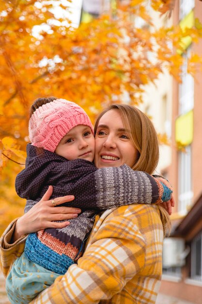 Portrait of happy and beautiful mother with her daughter in her arms among autumn leaves on city str...
